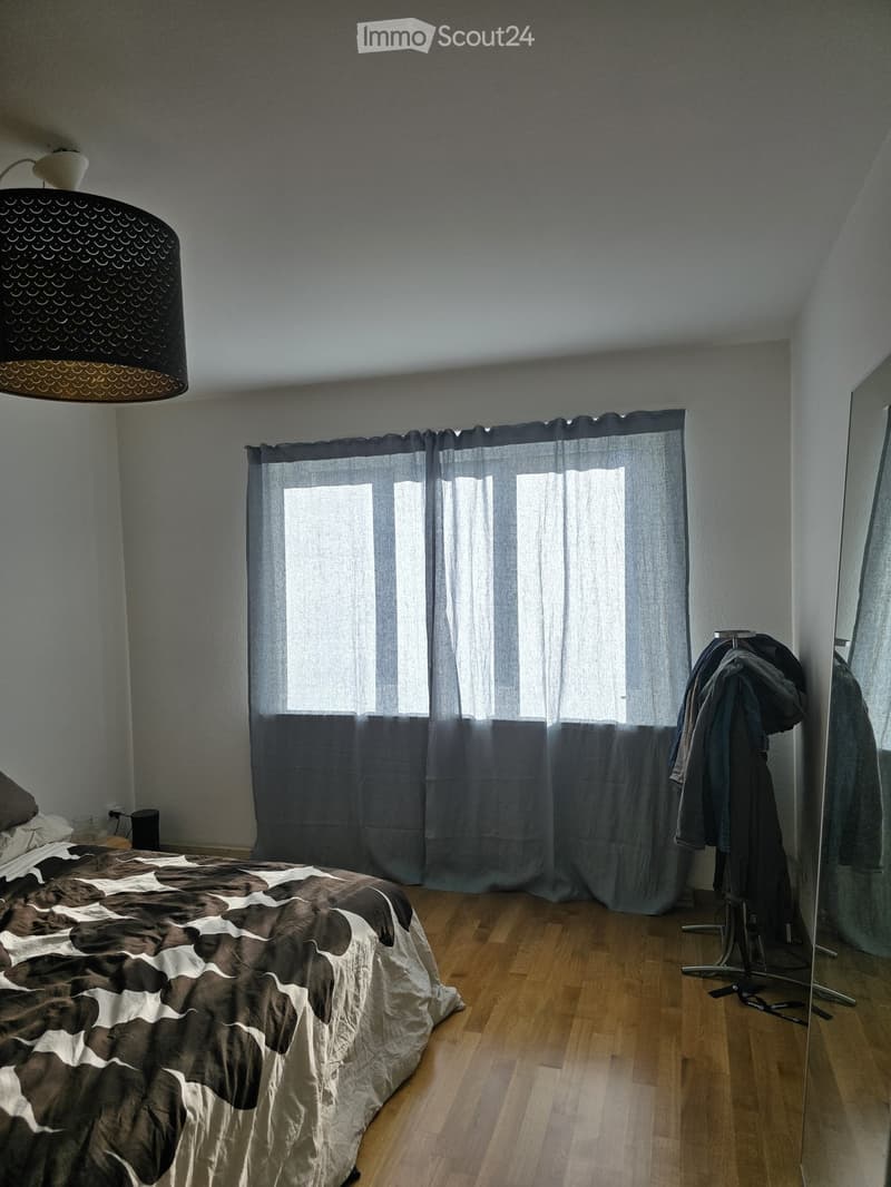 A modern bedroom with wooden flooring, a large window with grey curtains, a bed with a brown and white duvet, a wall-mounted mirror, and a clothing rack with coats.