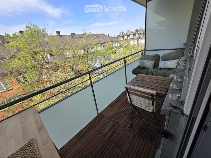 Balcony with wooden decking, glass railing, sofa, table, and chair, overlooking a cityscape with trees and rooftops.