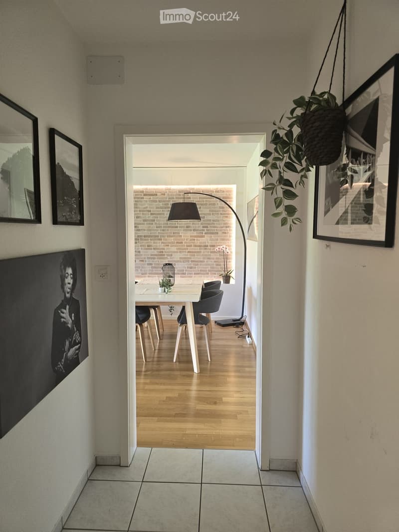 Hallway with tiled floor, leading to a dining area with a wooden table, chairs, and a floor lamp. Artwork is displayed on the walls.
