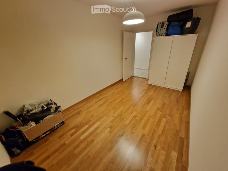 Empty room with wooden flooring, white walls, a white wardrobe, and a hanging light fixture. There are some boxes and bags placed near the walls.