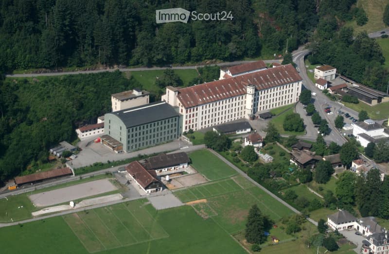 Aerial view of a large industrial or institutional complex with multiple buildings, surrounded by green fields and forested areas.