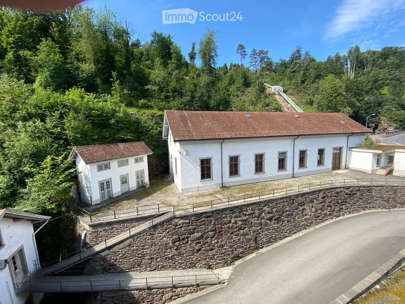 Aerial view of a white building with a brown tiled roof, surrounded by greenery and a stone retaining wall.