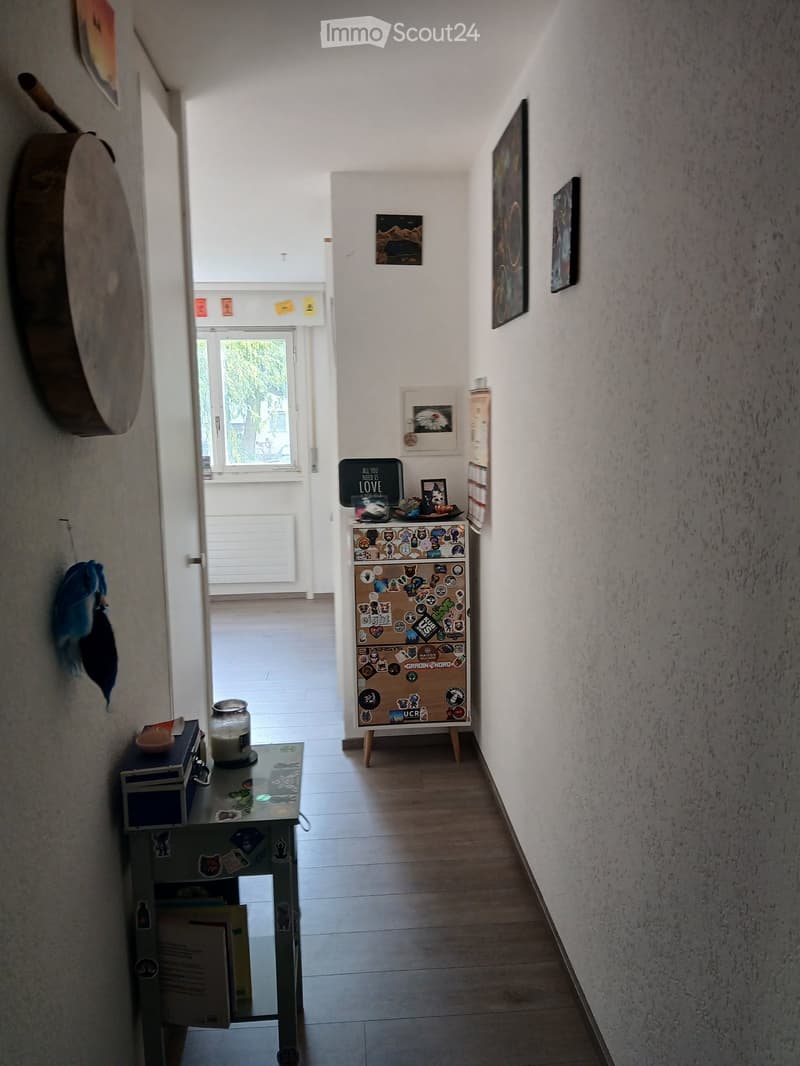 A narrow hallway with wooden flooring, white walls, and several pieces of art and decorations including a small table with items, a shelf with stickers, and framed pictures.
