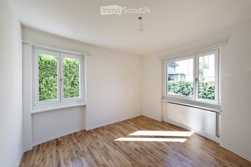 Empty room with light wooden flooring, white walls, and large windows offering views of greenery outside. A radiator is visible beneath one window.