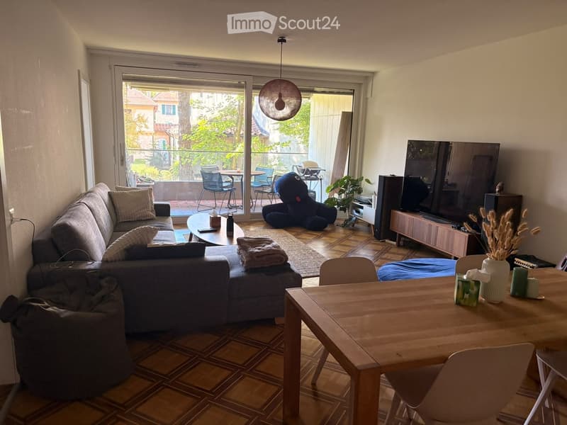 Modern living room with a grey sectional sofa, coffee table, dining table, chairs, TV, and a large window leading to a balcony.