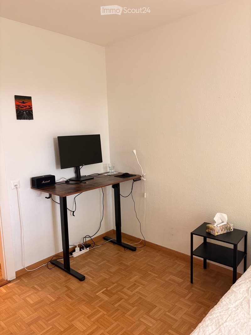 A modern, furnished room with a wooden herringbone floor, a standing desk with a monitor, a small black side table with a tissue box, and a partially visible bed with white bedding.