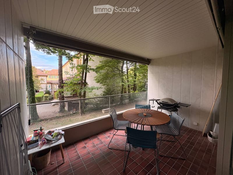 Balcony with red tiled floor, wooden table, four chairs, grill, and a small side table with items on it.