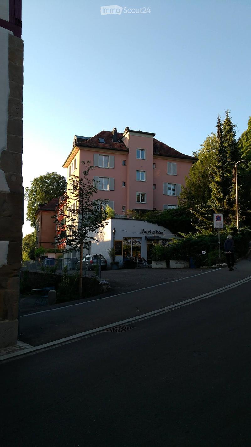 A multi-story residential building with pink facade and multiple windows, adjacent to a small shop with outdoor seating and a car parked nearby.