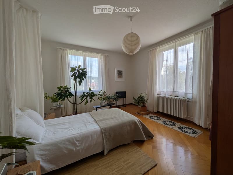 Bright bedroom with wooden flooring, large windows, a bed with white bedding, nightstand, small table, and potted plants.