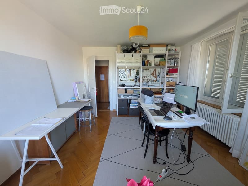 A well-lit, organized workspace with wooden flooring, two desks, chairs, a laptop, a large screen, shelves filled with books and supplies, and a small rug.