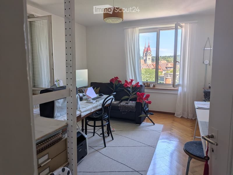 Bright living room with wooden flooring, large windows offering a cityscape view, a sofa, a laptop on a table, and a flower vase on the floor.