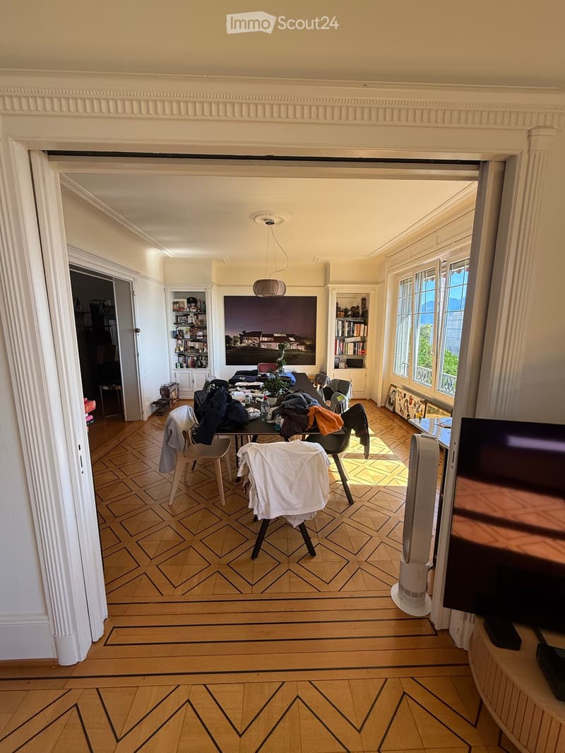 A spacious living room with wooden herringbone flooring, large windows, a TV screen, a fan, a table with chairs, and a bookshelf with books and decorative items.