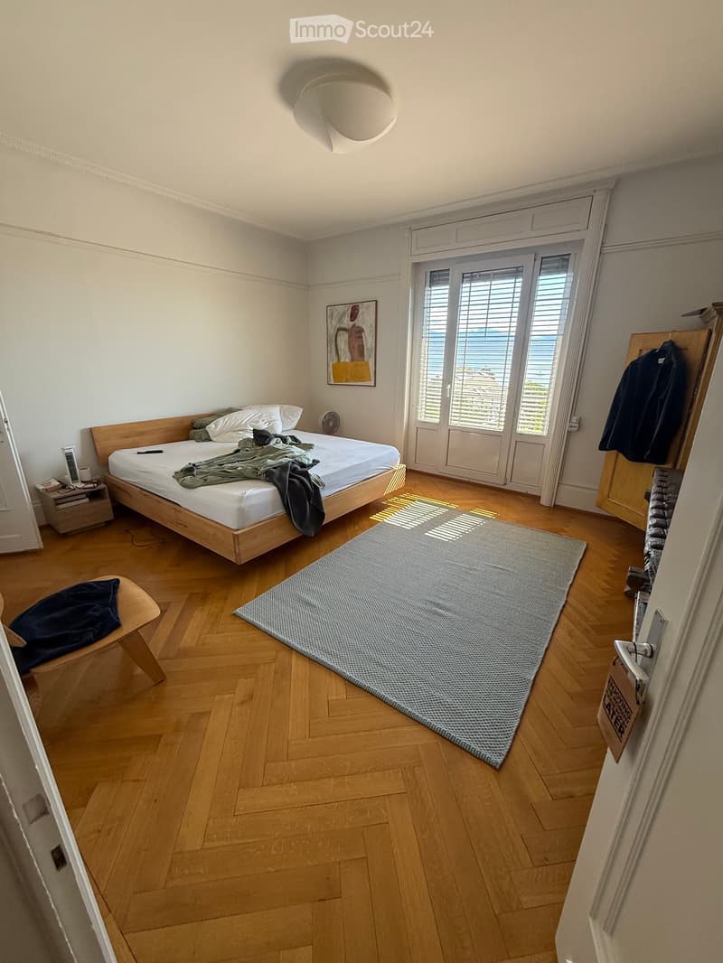 Modern bedroom with wooden flooring, a large bed with white linens, a nightstand, a chair, a rug, and a painting on the wall. Large windows with blinds provide natural light.