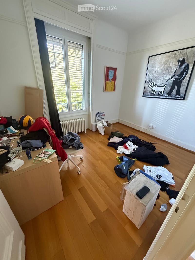 Empty room with wooden flooring, white walls, large windows with blinds, and some scattered items including clothes, a ball, and a few boxes.