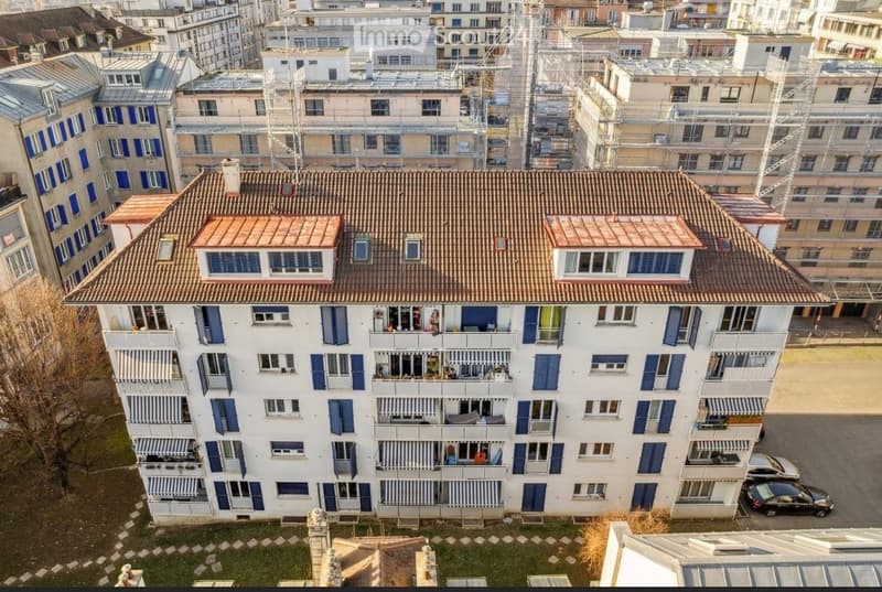 A multi-story residential building with white walls, blue shutters, and multiple balconies. The roof is tiled, and the surrounding area includes other buildings and parked cars.