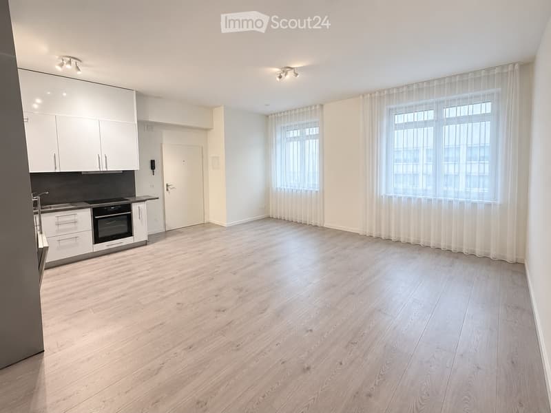 Empty room with light wooden flooring, white walls, large windows with sheer curtains, and a modern kitchen area with white cabinets and stainless steel appliances.