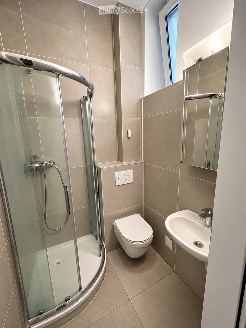 Modern bathroom with beige tiled walls and floor, featuring a corner shower with glass enclosure, a toilet, a sink with a modern faucet, and a mirror above the sink.