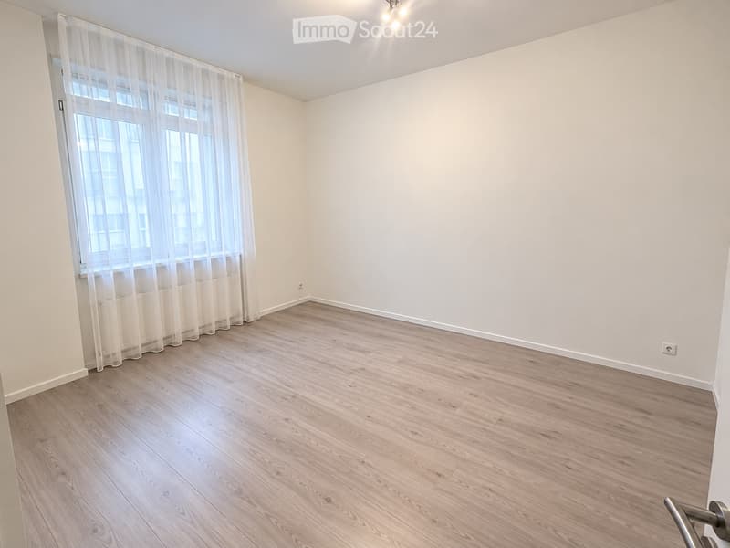 Empty room with light wooden flooring, white walls, and a large window with sheer curtains.