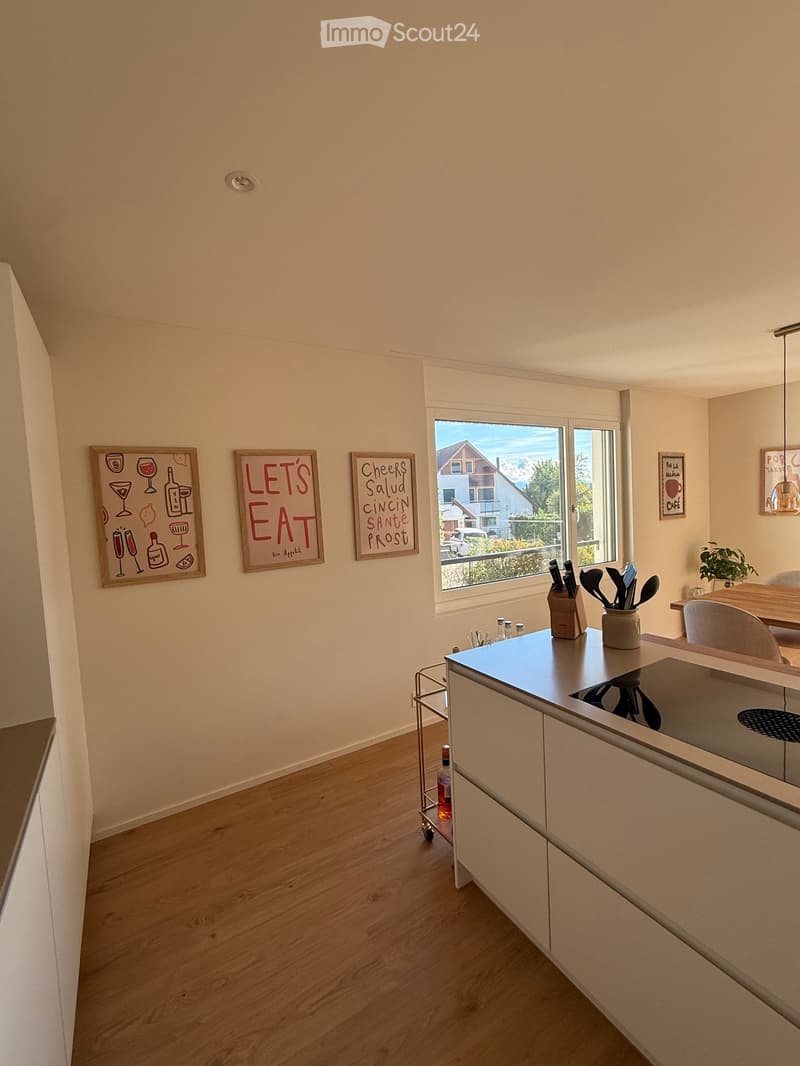 Modern kitchen with light wood flooring, white cabinets, and a central island. Features a knife block, a small cart with a bottle, and framed artwork on the walls.
