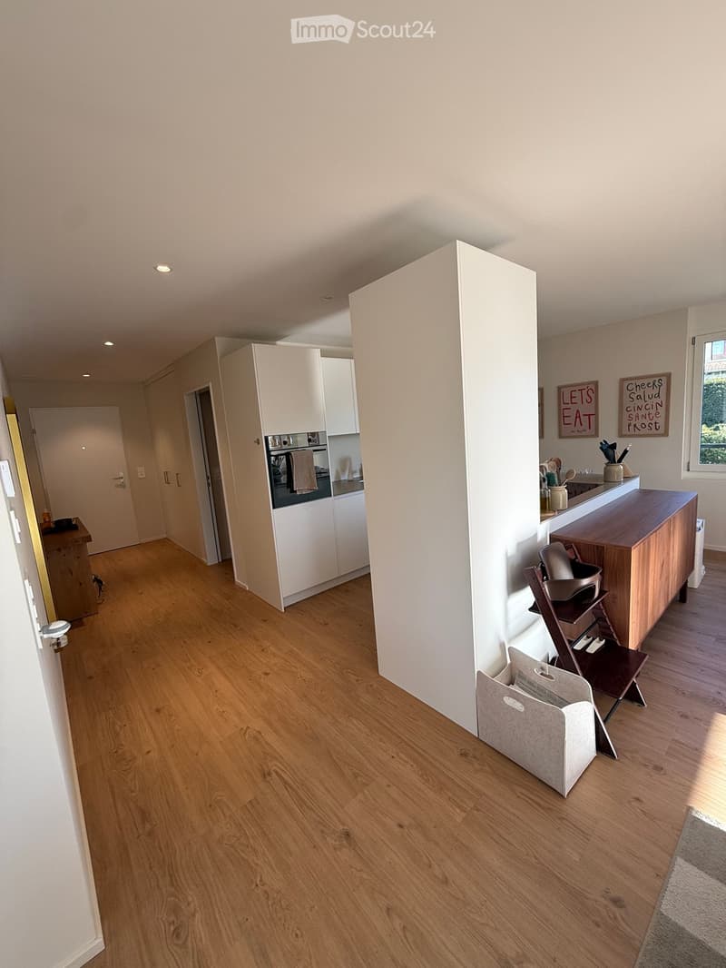 Open-plan living and dining area with light wooden flooring, white walls, and modern white cabinets. Features a baby chair, a counter with a framed sign, and an oven in the background.