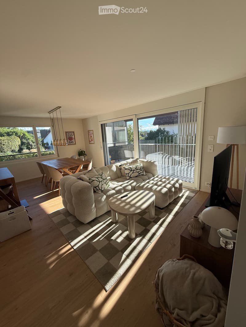 Bright living room with a large sectional sofa, coffee table, dining area, and large windows leading to a balcony.