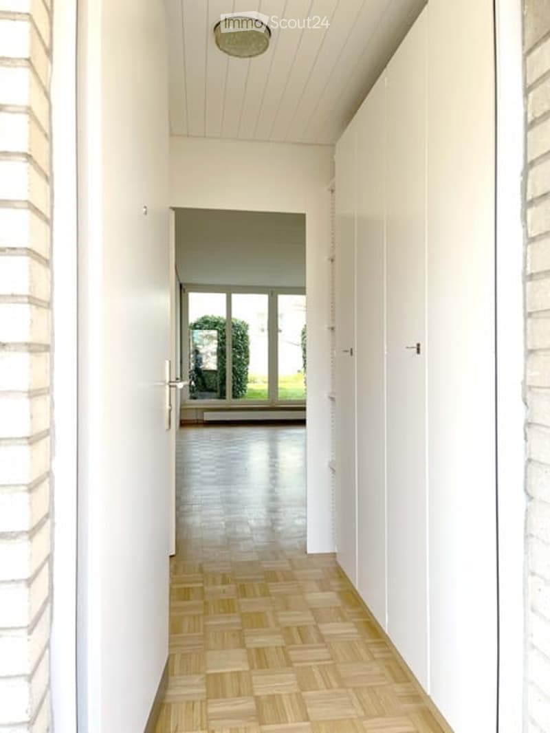 Bright hallway with wooden flooring, white walls, and a ceiling light fixture. The hallway leads to a room with large windows and a view of greenery outside.