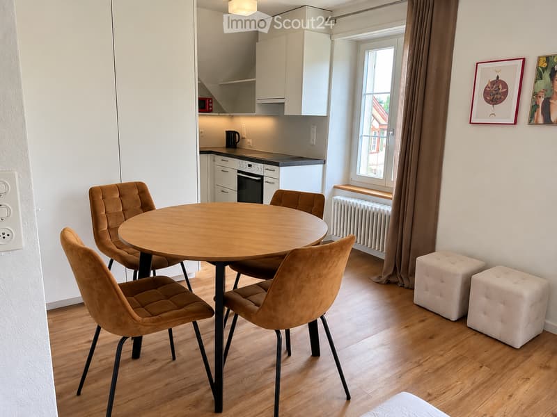 A modern, well-lit room with a round wooden dining table surrounded by four brown upholstered chairs, two white tufted stools, white kitchen cabinets, an oven, microwave, and a window with brown curtains.