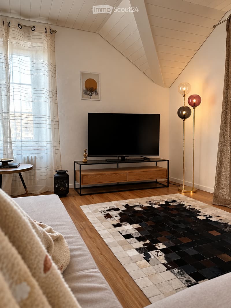 Modern living room with a slanted ceiling, wooden flooring, a large television on a wooden and black stand, a patterned rug, a round side table, and floor lamps.