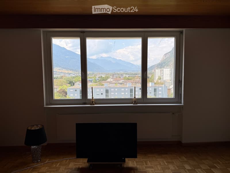 Empty room with wooden flooring, large windows offering a scenic mountain view, a table lamp, and a television on a stand.