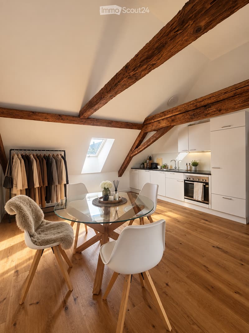 Bright, airy attic space with exposed wooden beams, white kitchen cabinets, a glass dining table with white chairs, and a furry throw on one chair. Features a skylight window and a coat rack with hanging coats.