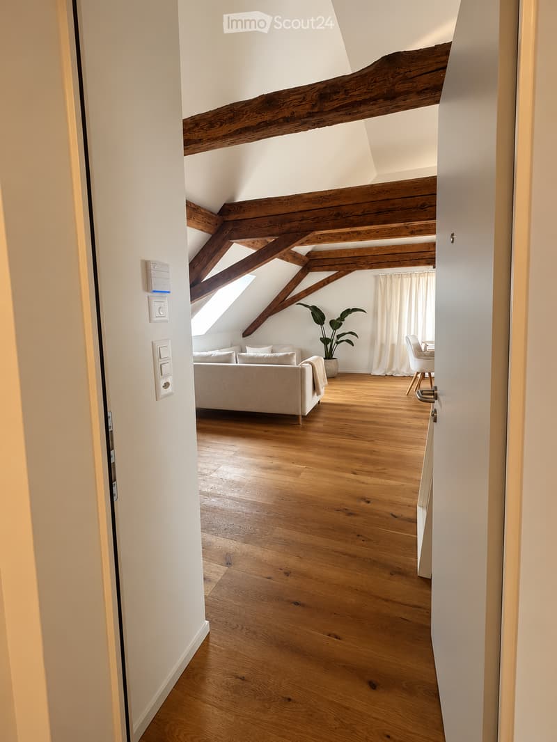 Bright living room with wooden floors, exposed wooden beams, a sofa, a chair, a small table, and a potted plant near a large window with white curtains.