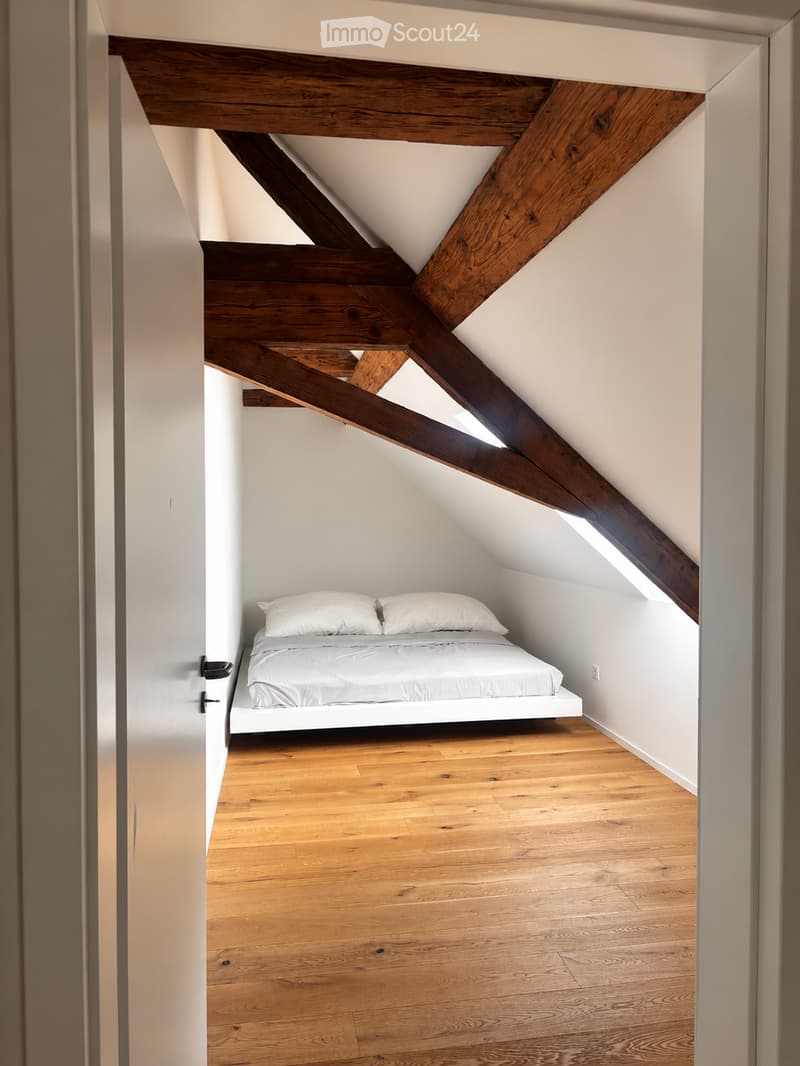 Cozy attic bedroom with exposed wooden beams, a white bed with two pillows, and wooden flooring.
