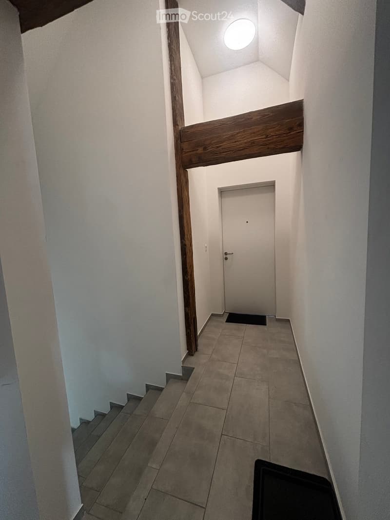 A narrow hallway with white walls, tiled floor, and a wooden staircase leading upwards. A closed door is visible at the end of the hallway.