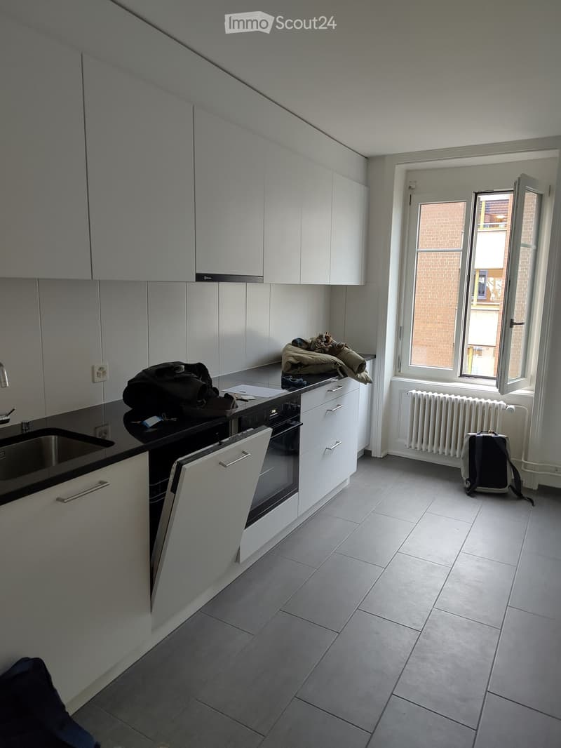 Modern kitchen with white cabinets, stainless steel sink, oven, and radiator. Personal items like a backpack and jacket are placed on the counter and floor.
