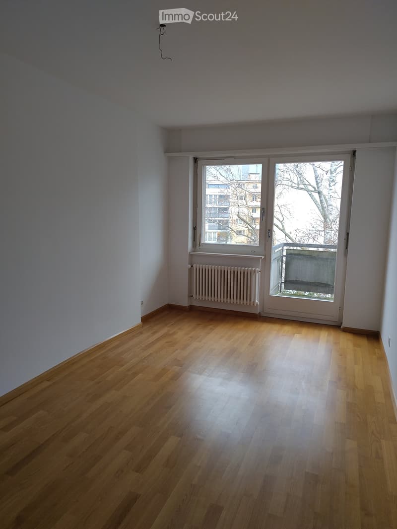 Empty room with wooden flooring, white walls, a radiator, and large windows with a view of the outside.