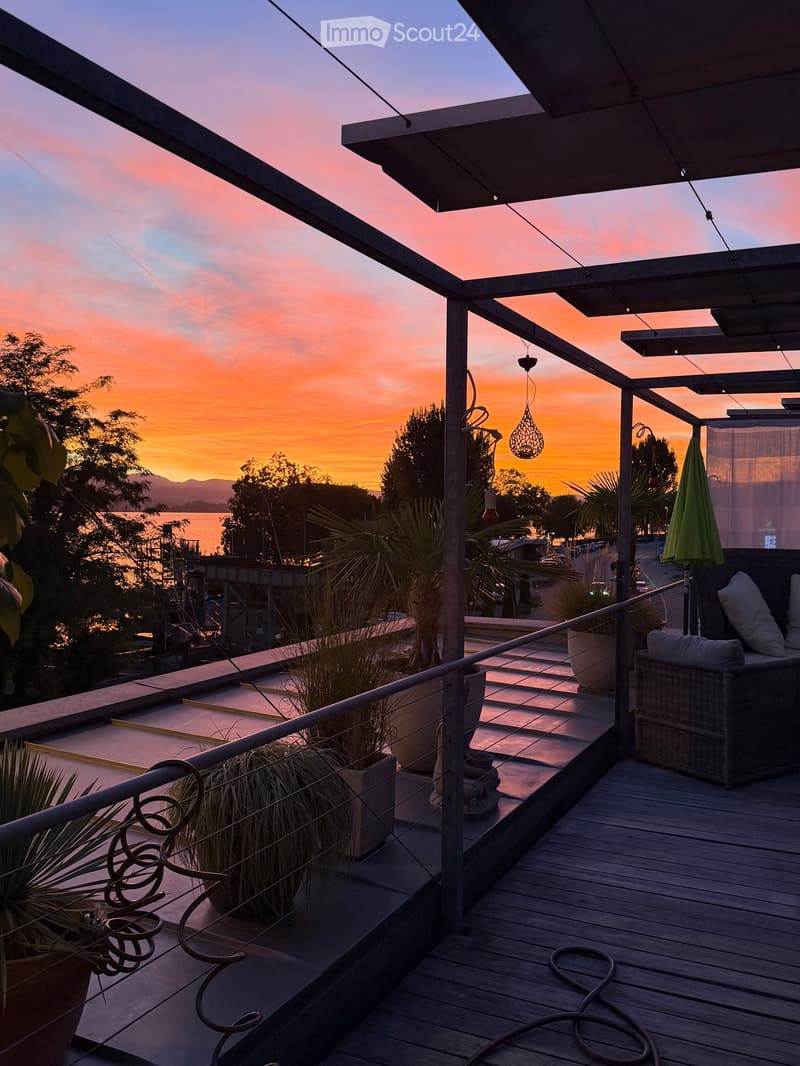 Wooden deck with a view of the sunset over the water, featuring a wicker sofa, potted plants, and an umbrella.
