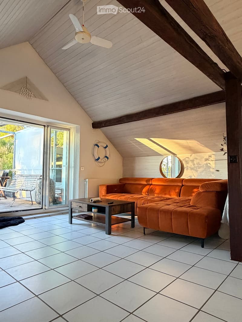Bright living room with white tiled flooring, orange sectional sofa, coffee table, and a round mirror on the wall. Large sliding glass doors lead to an outdoor area.
