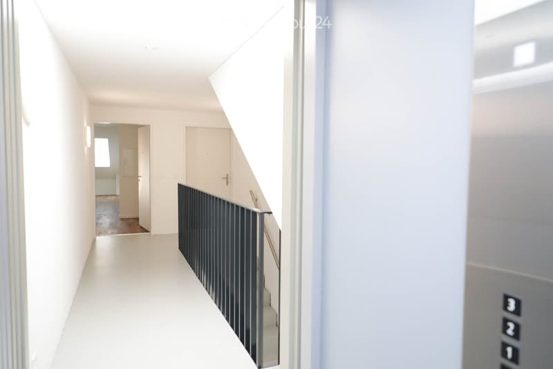 Modern hallway with white walls, light-colored flooring, and a staircase with a black metal railing. An elevator door is visible on the right side.