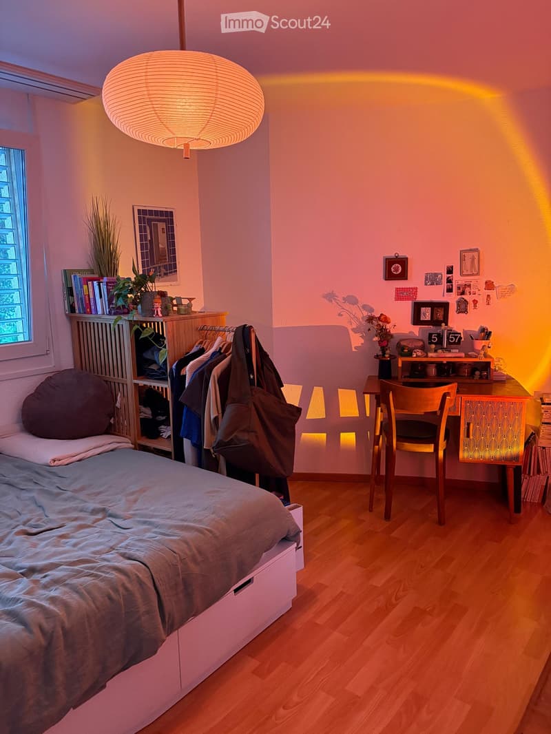 Cozy bedroom with a bed, desk, chair, and a hanging lamp. The room features wooden flooring, a window with a view, and various decorative items including books, plants, and artwork.