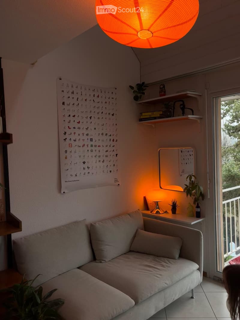 Cozy living room with a white sofa, small table, shelves with books and plants, and a large window leading to a balcony.