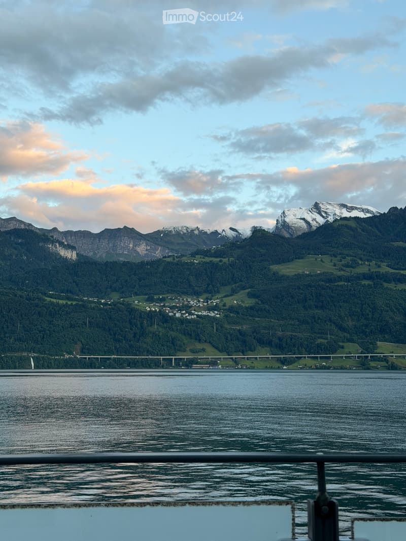 A scenic view of a lake with snow-capped mountains in the background, featuring a bridge and a small town nestled among the trees.