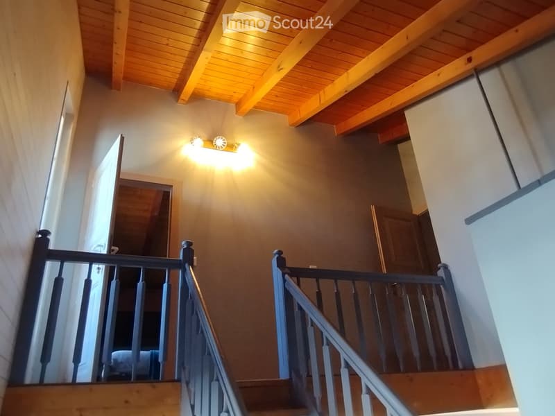 Staircase with wooden steps and dark railings, wooden ceiling beams, warm wall lighting, and open doorways leading to other rooms.