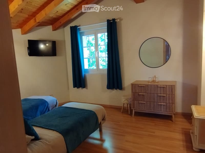 Cozy bedroom with wooden floors, two single beds, a dresser, a round mirror, and a television mounted on the wall.