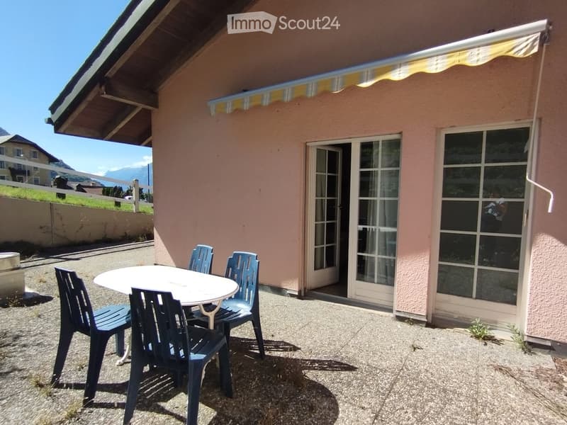 Balcony with a table and four chairs, attached to a pink house with large windows and a covered awning.