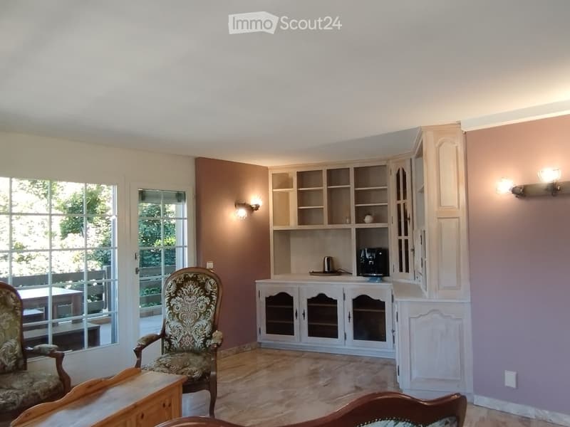 A well-lit room with wooden flooring, large windows, a patterned armchair, a small table, and a white cabinet with glass doors containing a coffee maker and other items.