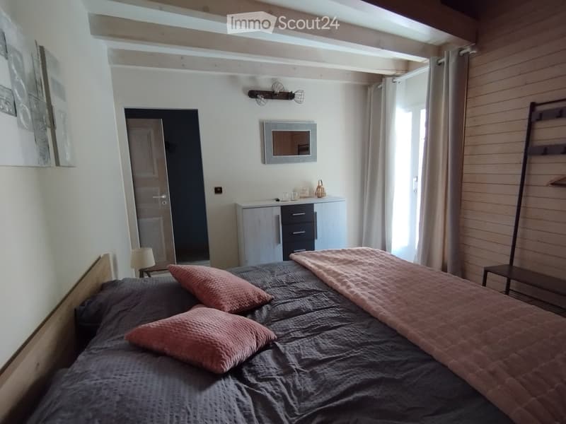 Cozy bedroom with a wooden bed, grey bedding, pink pillows, a nightstand with a lamp, a cabinet with drawers, a mirror, and a wardrobe. The room features exposed wooden beams and large windows with curtains.