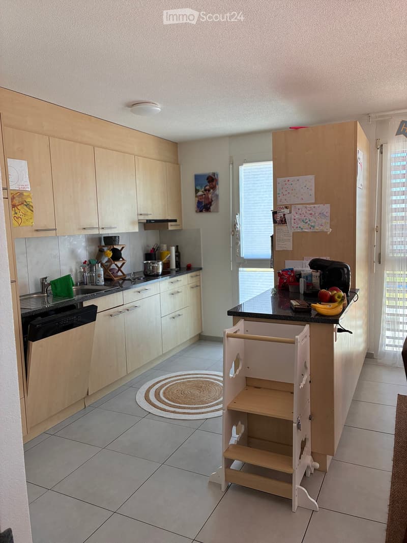 Modern kitchen with light wooden cabinets, black countertop, sink, dishwasher, and a small wooden stool for a child. There is a fruit bowl with apples and bananas on the counter, and a small round rug on the floor.