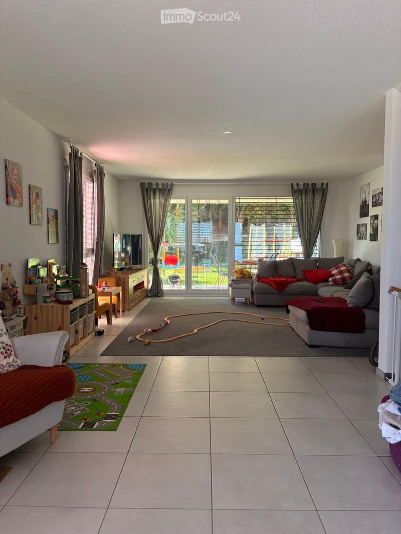 Living room with grey sectional sofa, ottoman, coffee table, TV, and toys on the floor. Large windows with curtains lead to a balcony.