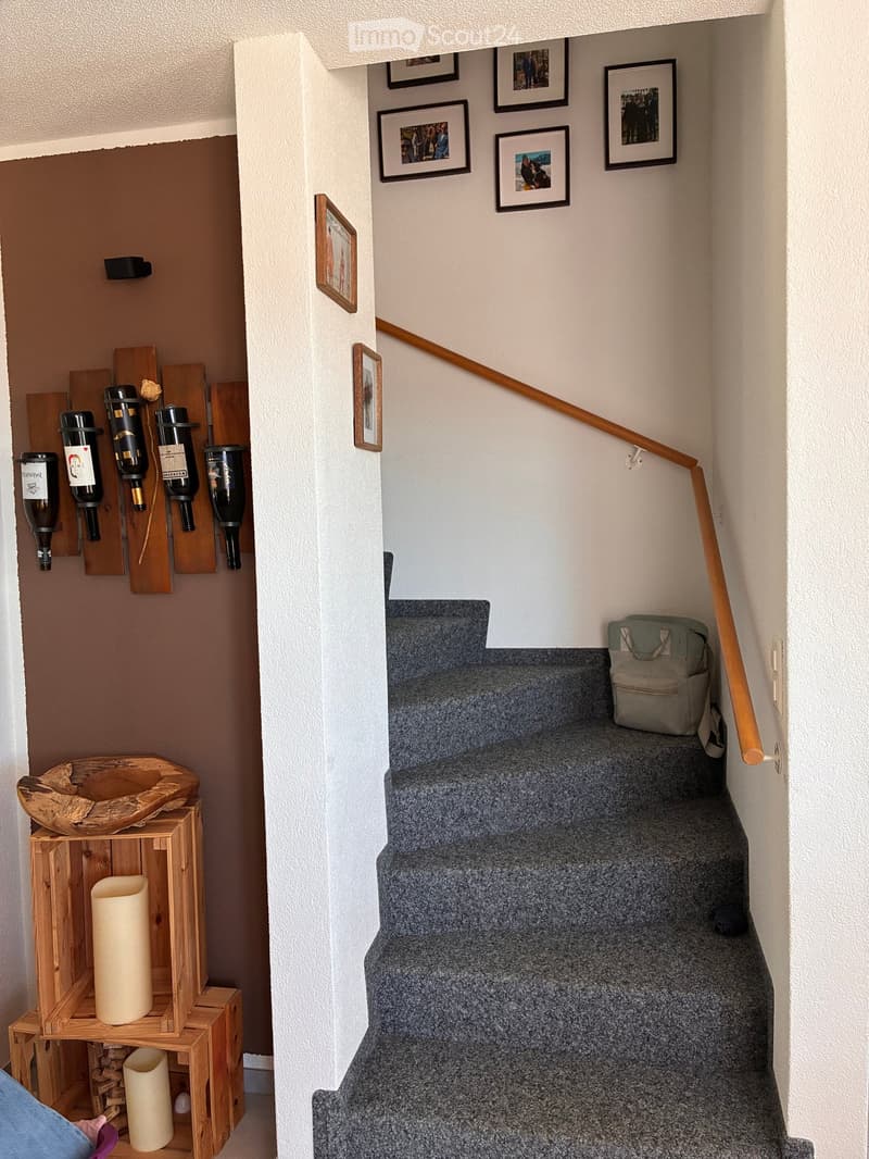 Staircase with wooden handrail, carpeted steps, and a small wooden shelf with decorative items and wine bottles on the wall.