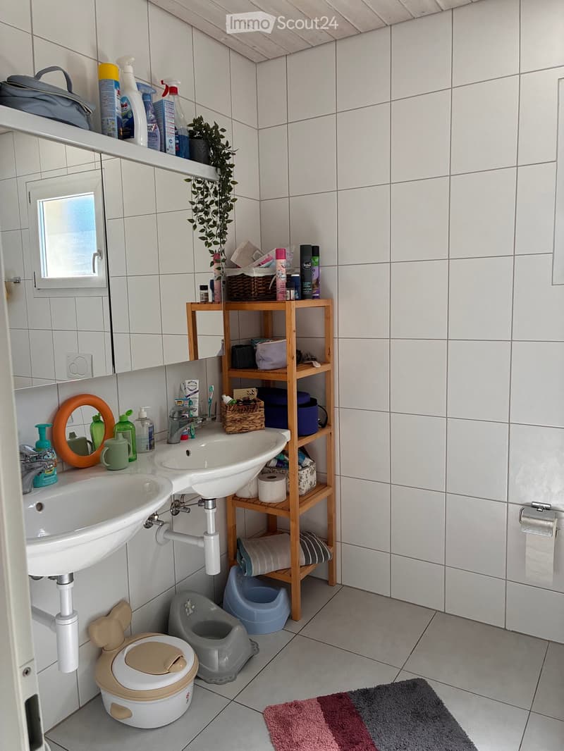 A small bathroom with white tiled walls and floor, featuring two sinks, a toilet, a wooden shelf with various items, and a small plant. There is also a toilet seat cover and a colorful bath mat on the floor.